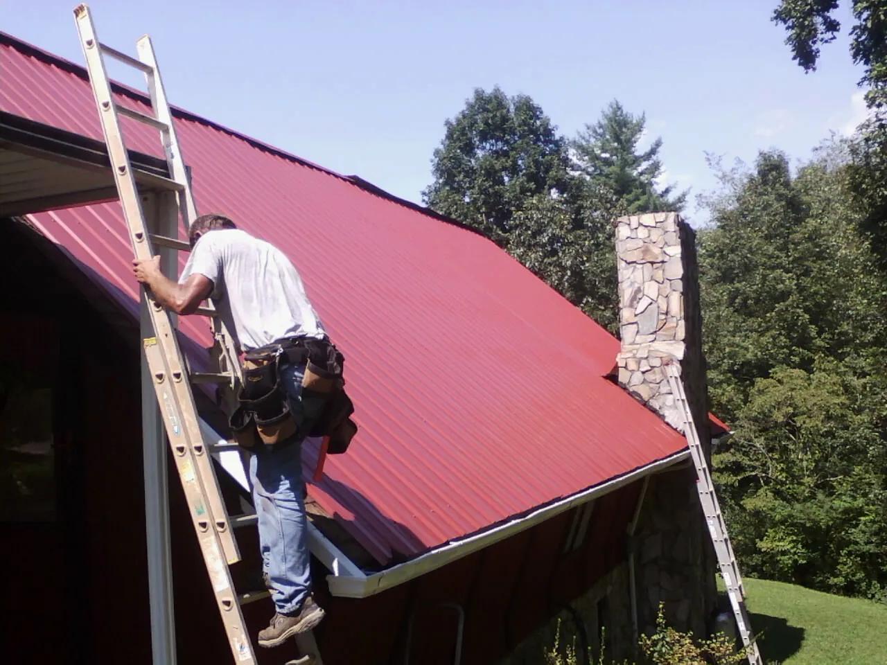 Roofer on a ladder beside a red metal roof and stone chimney — Soffit & Fascia Repair by Roofing Brothers in Stonyford